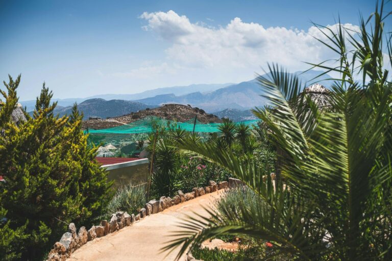 A picturesque pathway surrounded by lush greenery with mountains and blue sky in Rethymno, Greece.
