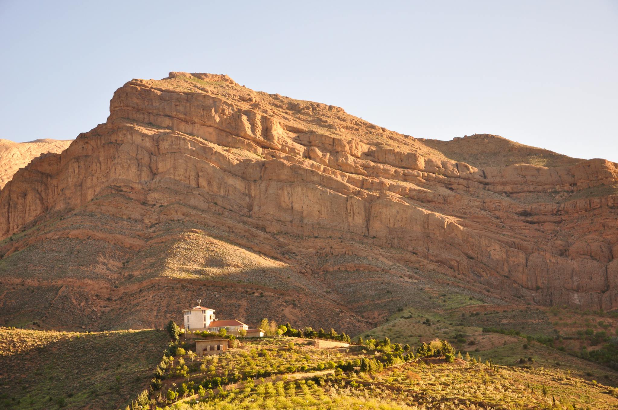 A secluded farmhouse sits peacefully at the base of a rugged mountain range, bathed in warm sunset light.