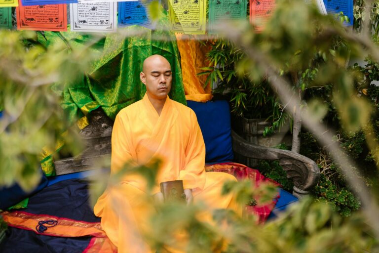 A serene monk in traditional attire meditates outdoors amidst colorful prayer flags and nature.